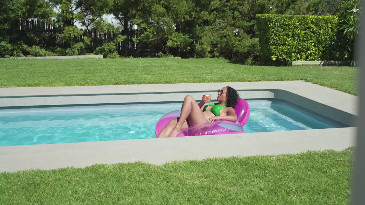 African American woman basking on pink float in yard pool, green suit holding cup, sipping, cooling