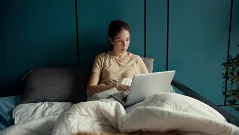 A-brunette-girl-lies-on-a-bed,-works-on-a-laptop-and-smiles-against-the-background-of-a-turquoise-wall-at-home.-Happy-working-remotely-from-home