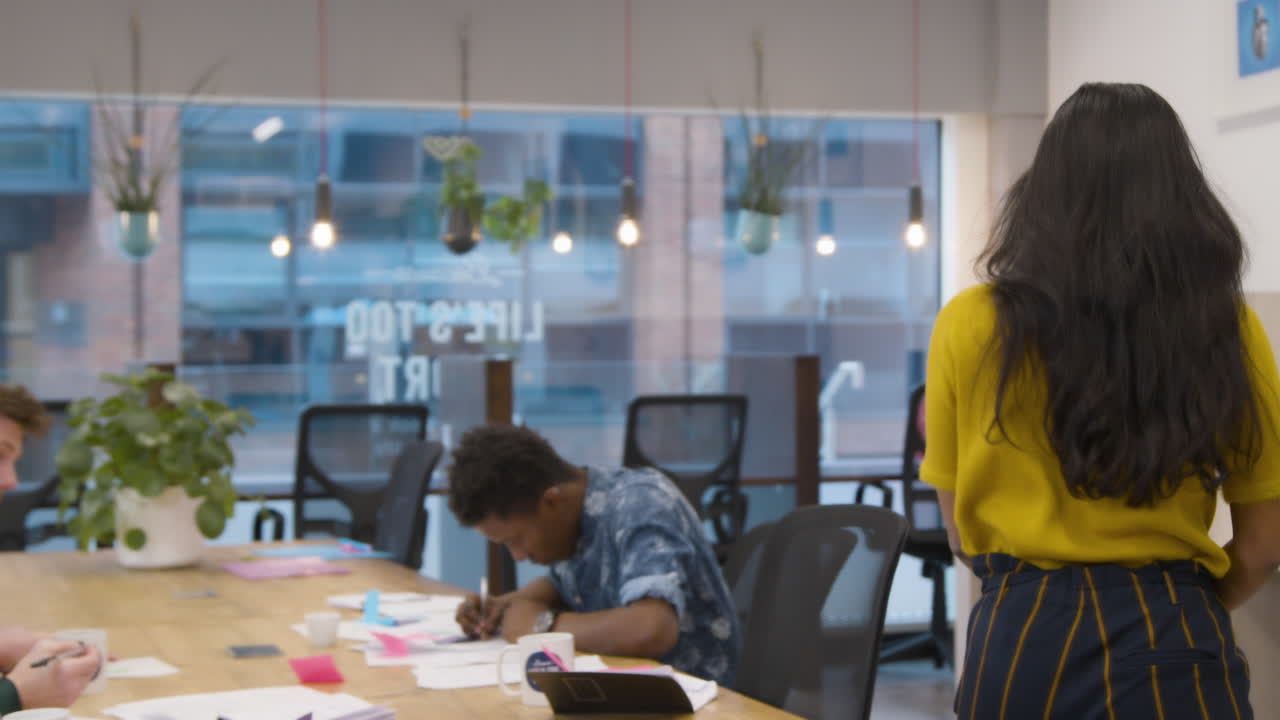 Woman Walking Into An Office And Sitting Down With Colleagues