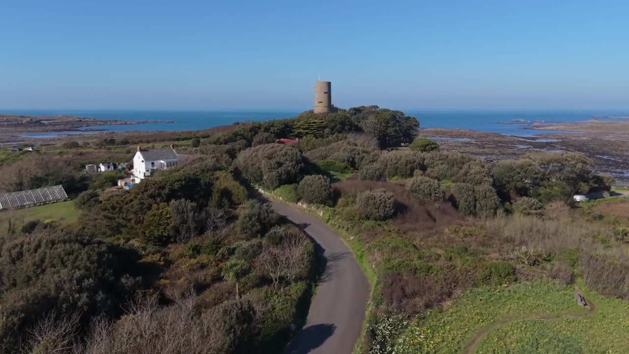 L’Eree, Guernsey: En landevei som fører til et gammelt tysk utkikkstårn på neset, med utsikt mot havet og Lihou-øya i den sene ettermiddagssolen under en klar blå himmel.