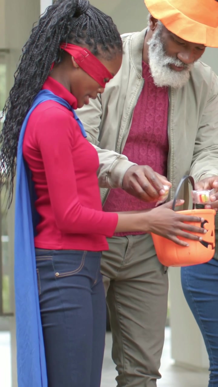 Vertical video: Steadying woman red-mask cape at home as man placing treats into pumpkin bucket
