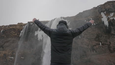 back of man walking in snow in warm winter clothes and raising arms in front of waterfall, slow motion