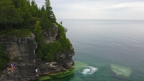 friends watch as young caucasian man leaps off cliff into clear lake