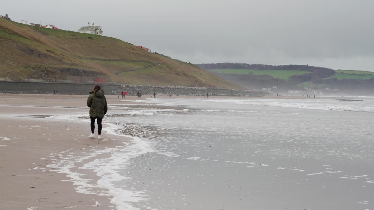 갈색 머리 소녀는 차가운 바람이 부는 오후에 조수가 들어오고 파도가 그녀에게 다가와서 그녀를 튀기기 위해 먼 곳에 있는 조용한 영국 해변을 따라 걷는다