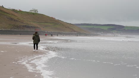 brunette girl walks along a quiet english beach in the distance on a cold windy afternoon as the tide comes in and waves approach her to splash her