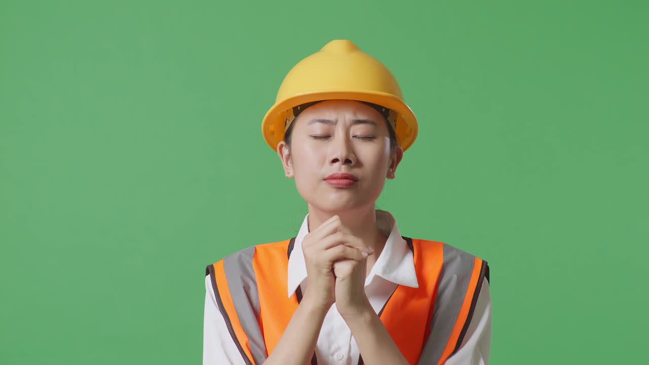 안전 헬을 입은 아시아 여성 엔지니어가 녹색 스크린 배경 스튜디오에 서 있는 동안 무언가를 위해 기도합니다.