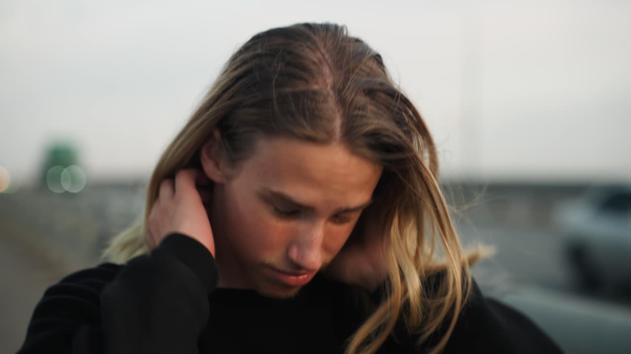 Retrato em close-up de um adolescente branco pensativo ajustando seu longo cabelo loiro em um viaduto urbano ao entardecer. Ele veste um moletom preto e tem uma mochila pendurada. Um vento suave e uma expressão pensativa são capturados nesta cena com iluminação dourada e um clima melancólico.