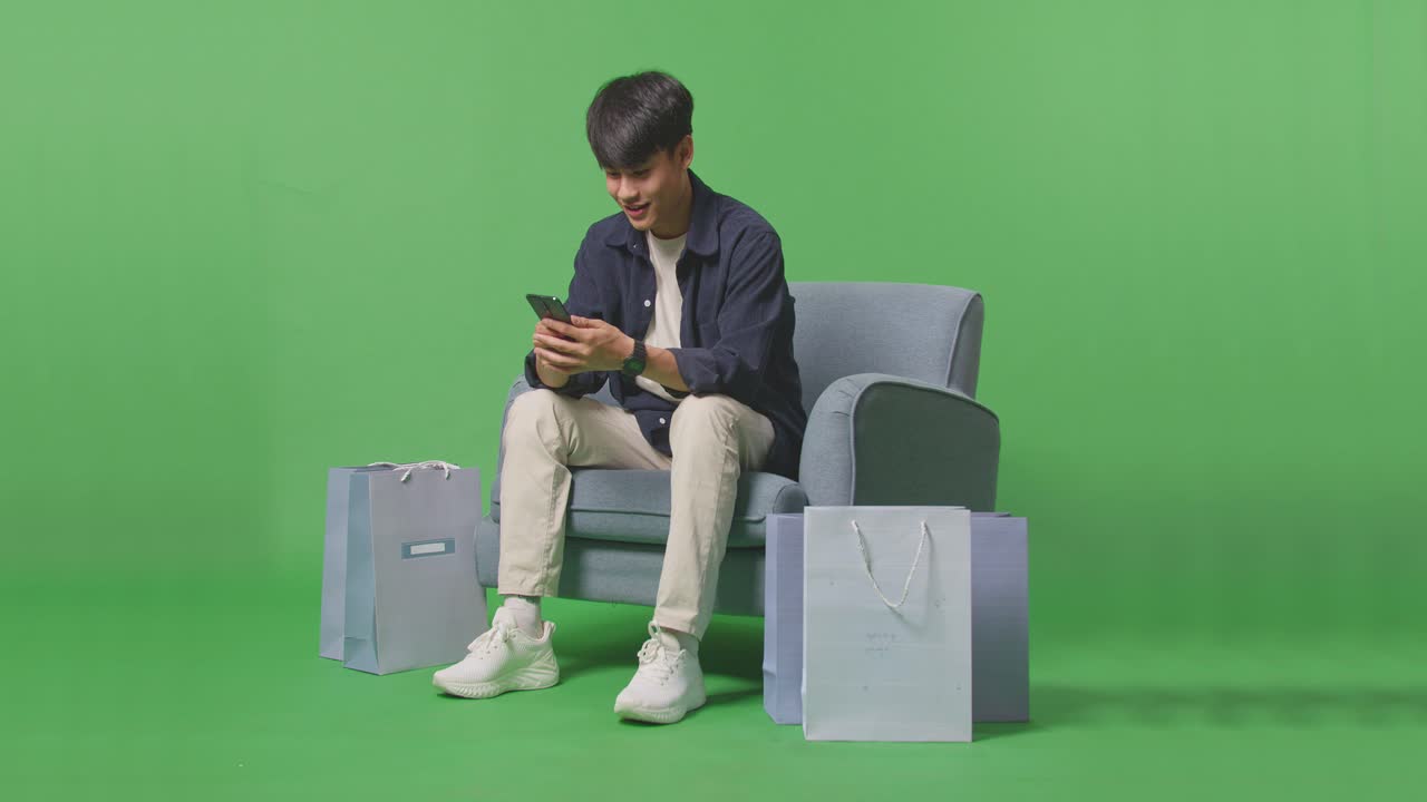 An Asian Shopping Man With Shopping Bag Enjoy Using Mobile Phone Shopping Online While Sitting On Sofa In Front Of Green Screen