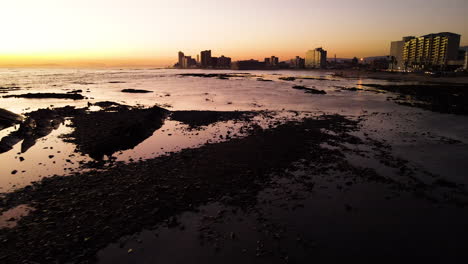 Flight-over-rocky-shallows-of-Strand-in-Western-Cape-at-sunset,-coastal-city