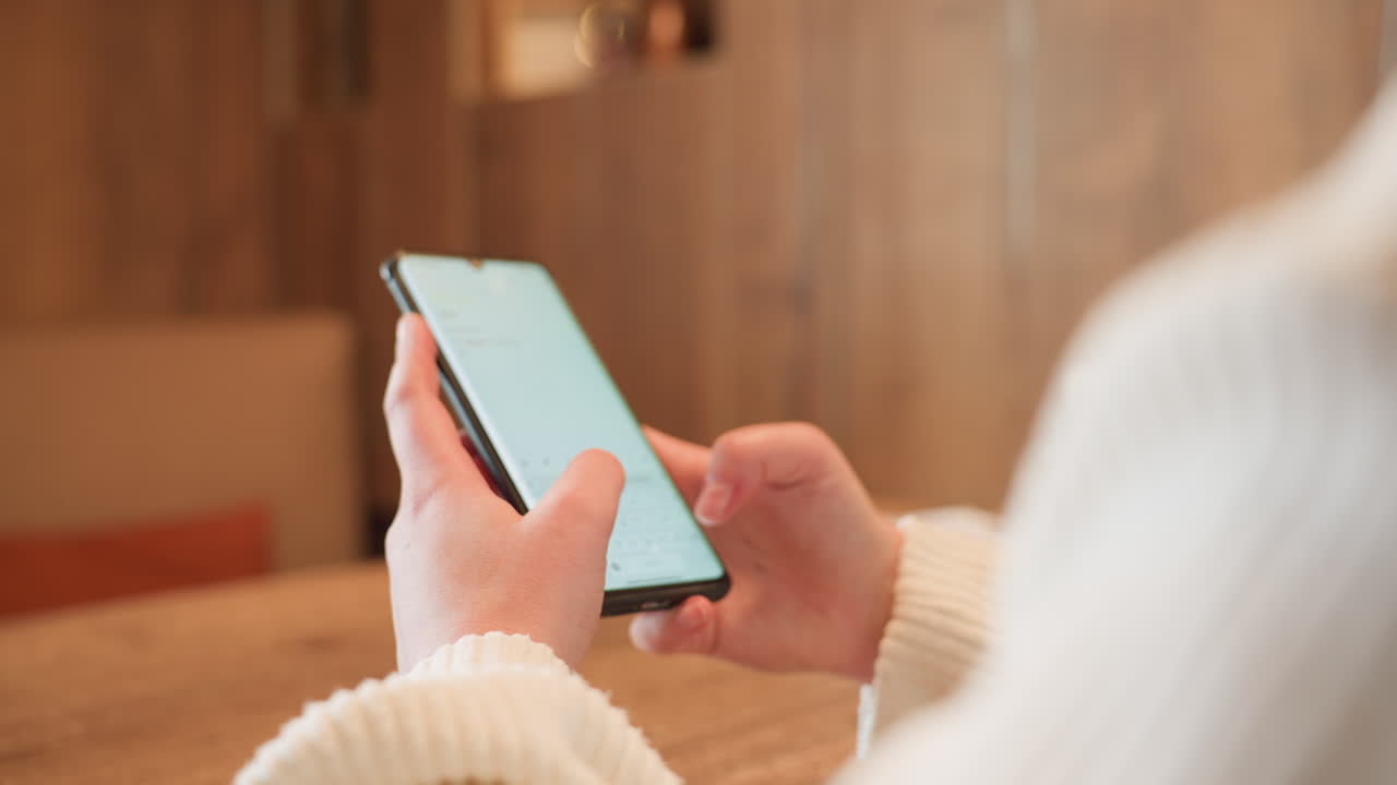 Close up hand view of woman using smartphone in warm wooden cafe interior, fingers actively engaged with screen, showcasing technology interaction in calm setting with blurred