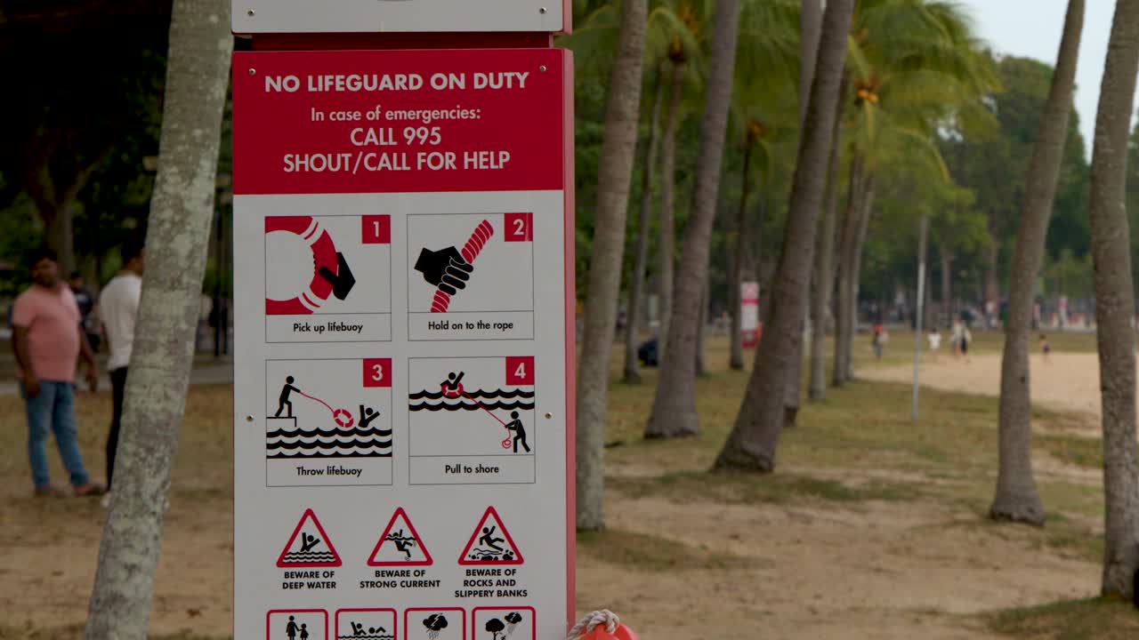 Un'inquadratura media statica cattura un cartello di avvertimento rosso del bagnino tra palme tropicali.