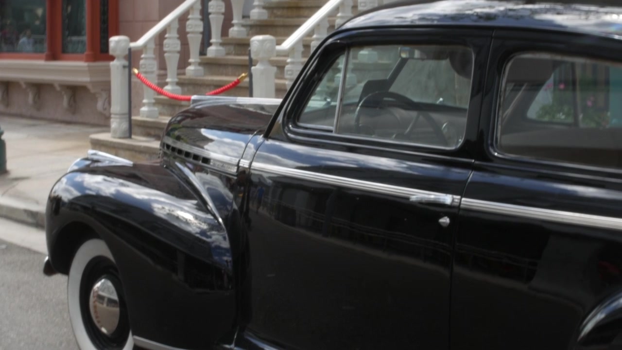 Coche aparcado junto a escalones de piedra y barreras de cuerda rojas en una calle tranquila de la ciudad. Primer plano de un sedán clásico negro con molduras cromadas y neumáticos de banda blanca.