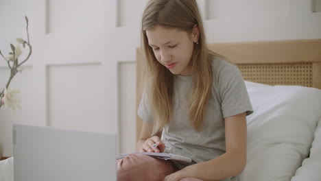 teen girl is preparing to online lesson by internet holding copybook sitting on bed in front of laptop