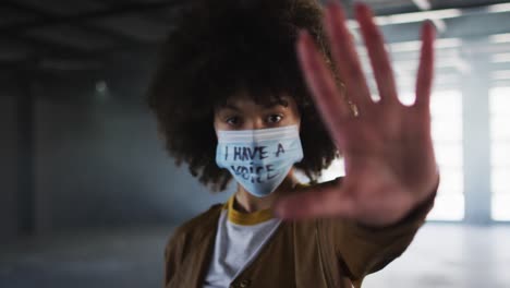 Portrait-of-african-american-woman-wearing-protest-face-mask-gesturing-stop-sign-in-empty-parking-ga