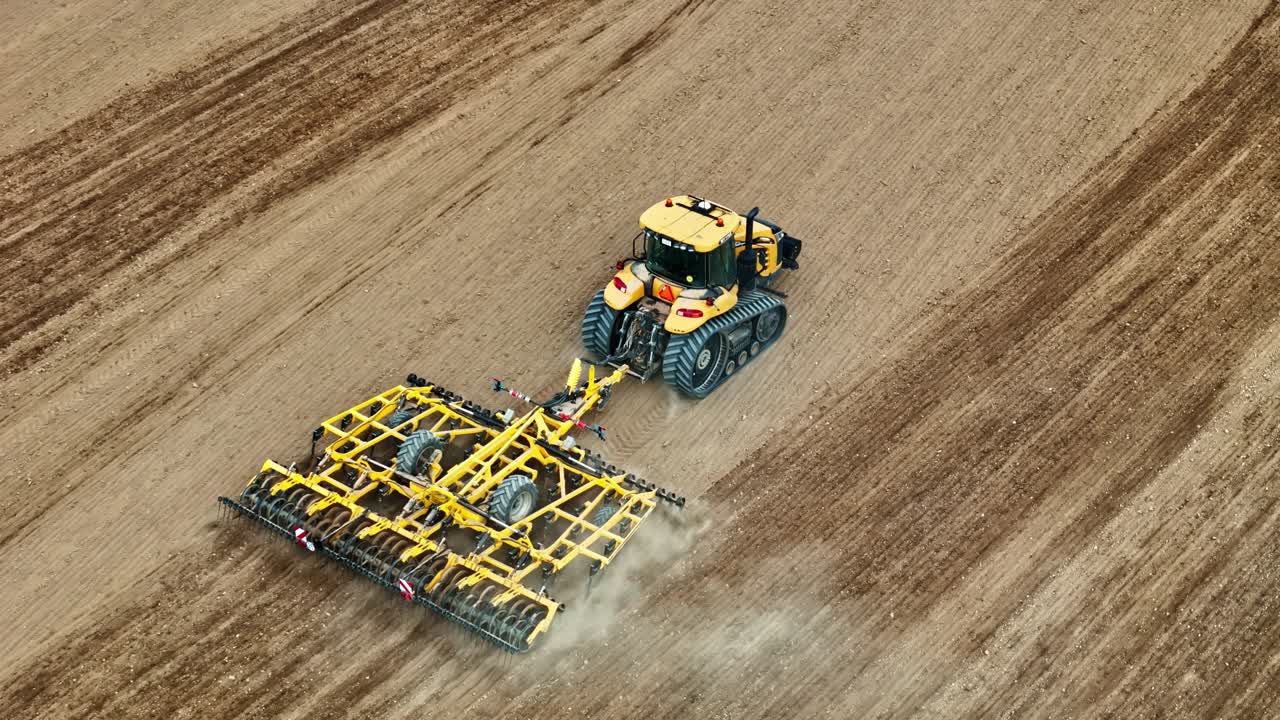 Droneoptagelse fra luften af en stor gul bæltegående traktor, der trækker en bred kultivator hen over en tør, åben mark. Støv stiger op bag redskabet, mens maskinen bearbejder den bare jord