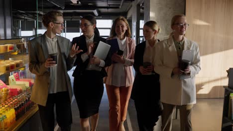 A-group-of-confident-businesswomen-in-business-clothes-walk-along-a-modern-office-during-a-meeting-with-coffee-in-their-hands-and-a-laptop