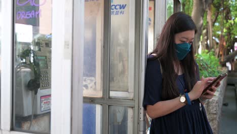 a woman texting and using a smartphone next to a public telephone booth, also called a payphone, as they are rapidly dying out and disappearing in the age of smartphones and the internet