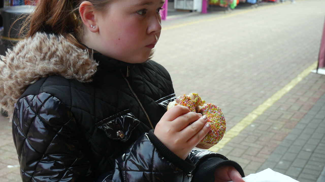 Una bambina felice con una giacca nera foderata di pelliccia che gusta una ciambella ricoperta di zuccherini in un mercato all'aperto.