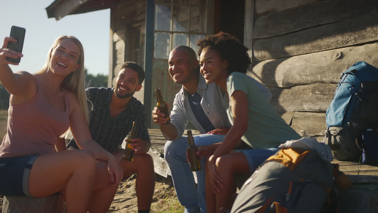 grupo de amigos de vacaciones sentados en el porche de una cabaña de campo bebiendo cerveza y tomando selfies