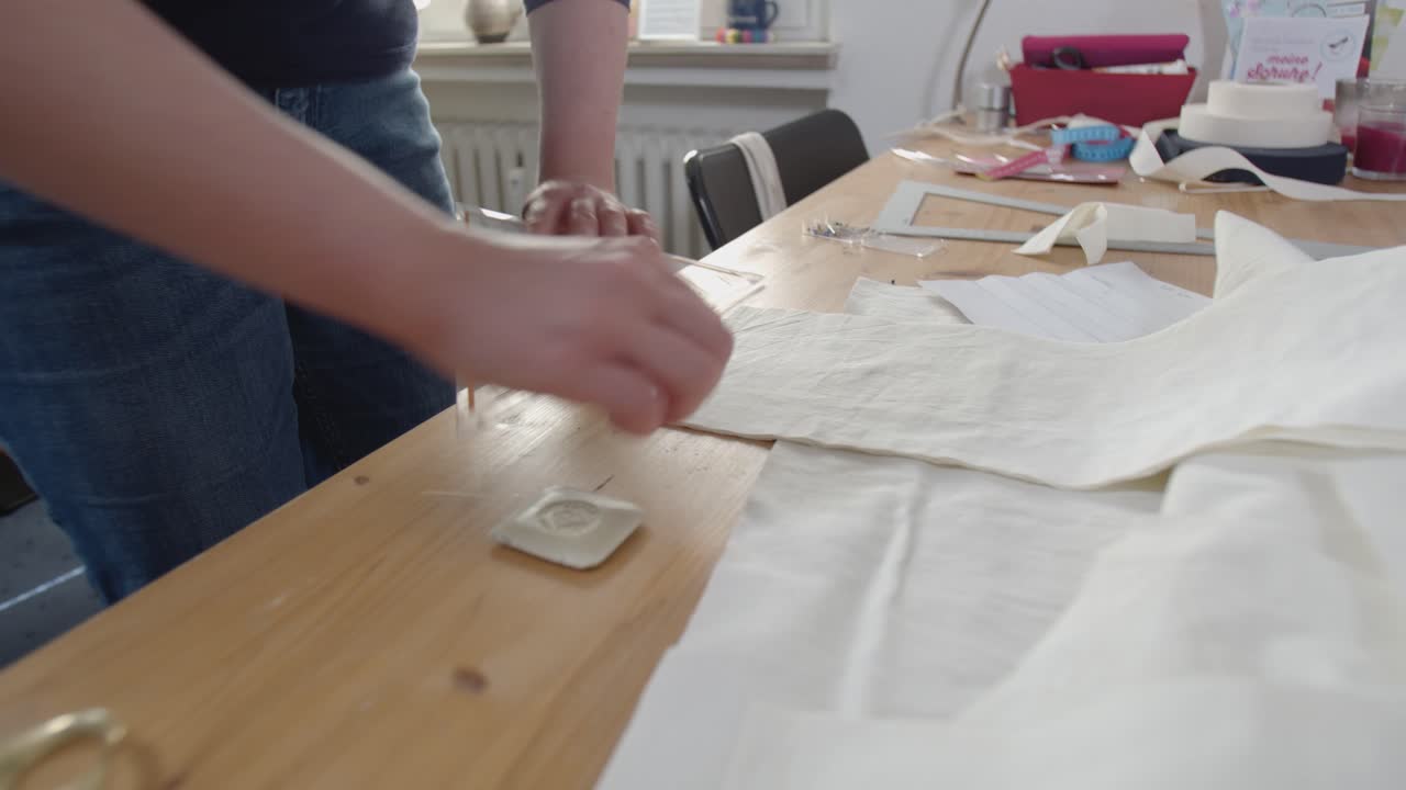A close-up view of seamstress hands marking out measurements on a white cloth on a table full of fabric and tools for sewing