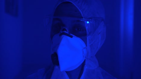 man wears protective suit, face mask and goggles, looks anxiously around dark misty room with flashing red blue light