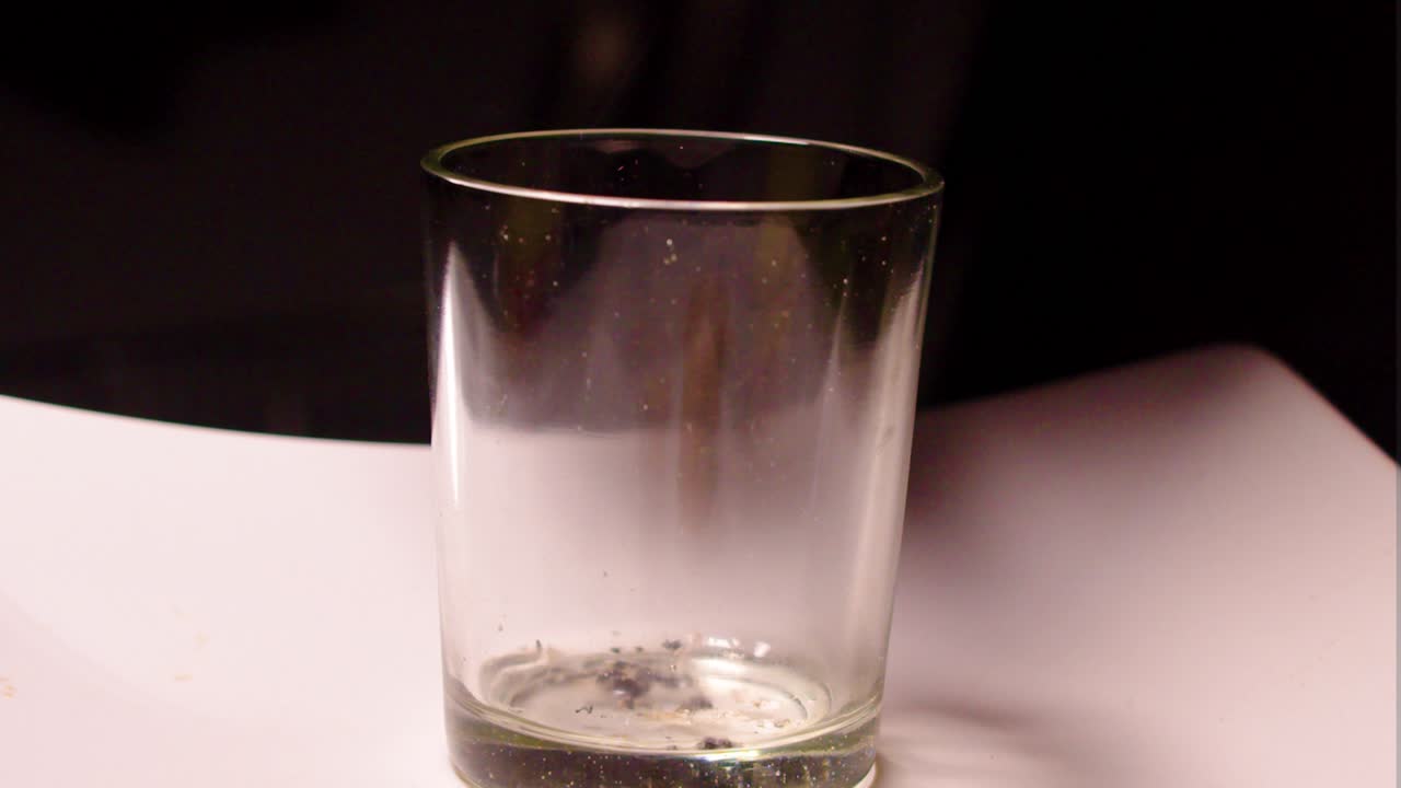 empty glass on white plate in the middle of the frame used as an ashtray for cigarette tobacco ash white Caucasian hand holding smoking product flicking remaining's medium close up lonely depression