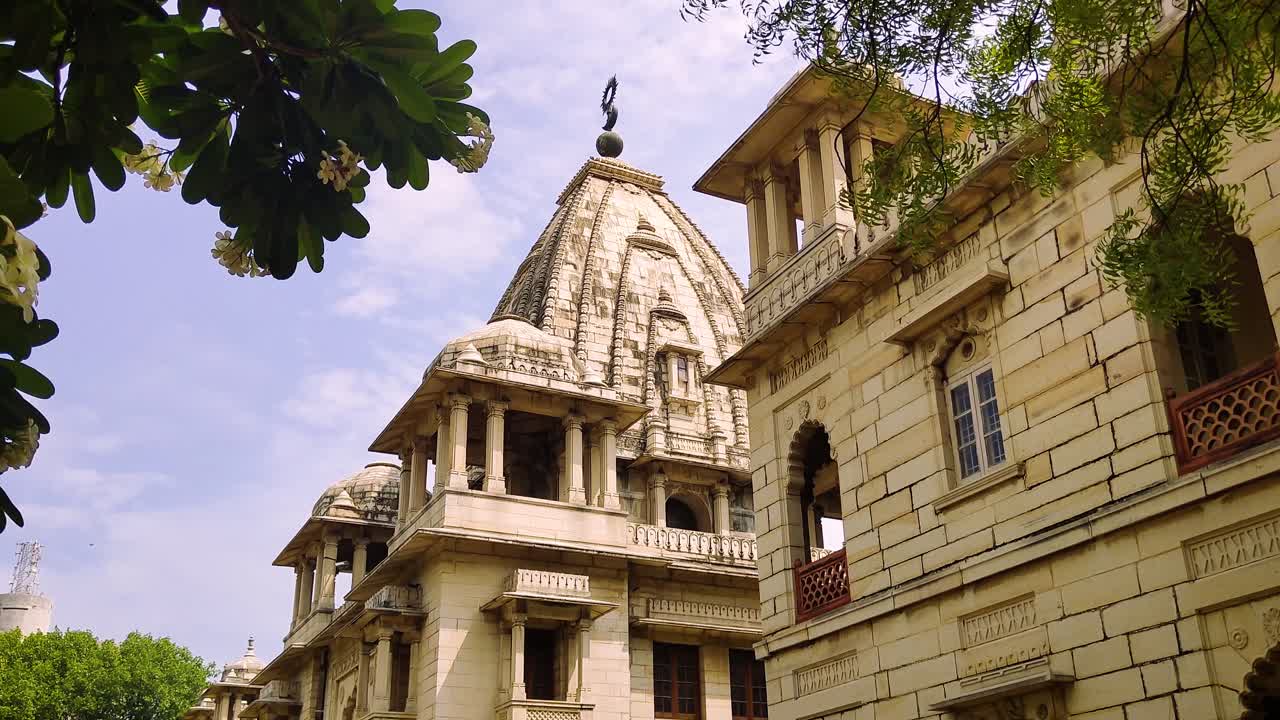 4K tilt-up video of Kirti Mandir temple in Vadodara, Gujarat, India. The shot showcases ornate heritage architecture against partly cloudy sky, framed by lush green trees