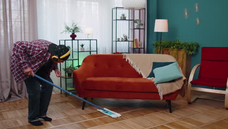 senior grandfather is mopping floor in light apartment living room at home, listening music, dancing