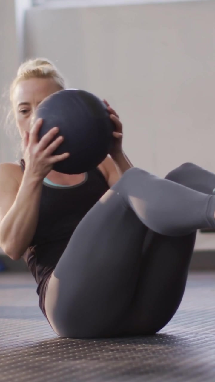 Vertical video: Woman doing Vsit on mat rotating medball for fitness with social overlays appearing