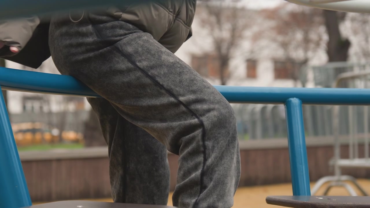 Young kid ascends metal railing. Young child gripping metal bar in calm determination during urban play. Small child in grey pants ascends blue railing with quiet resolve under cloudy sky