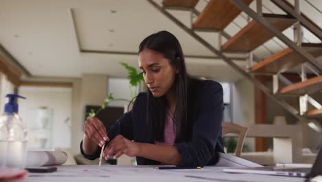 Mixed-race-woman-by-desk-at-home-using-laptop-looking-through-blueprints