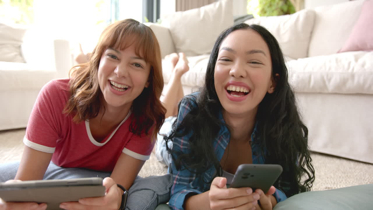 Two young biracial female friends lying on the floor smiling and holding smartphones