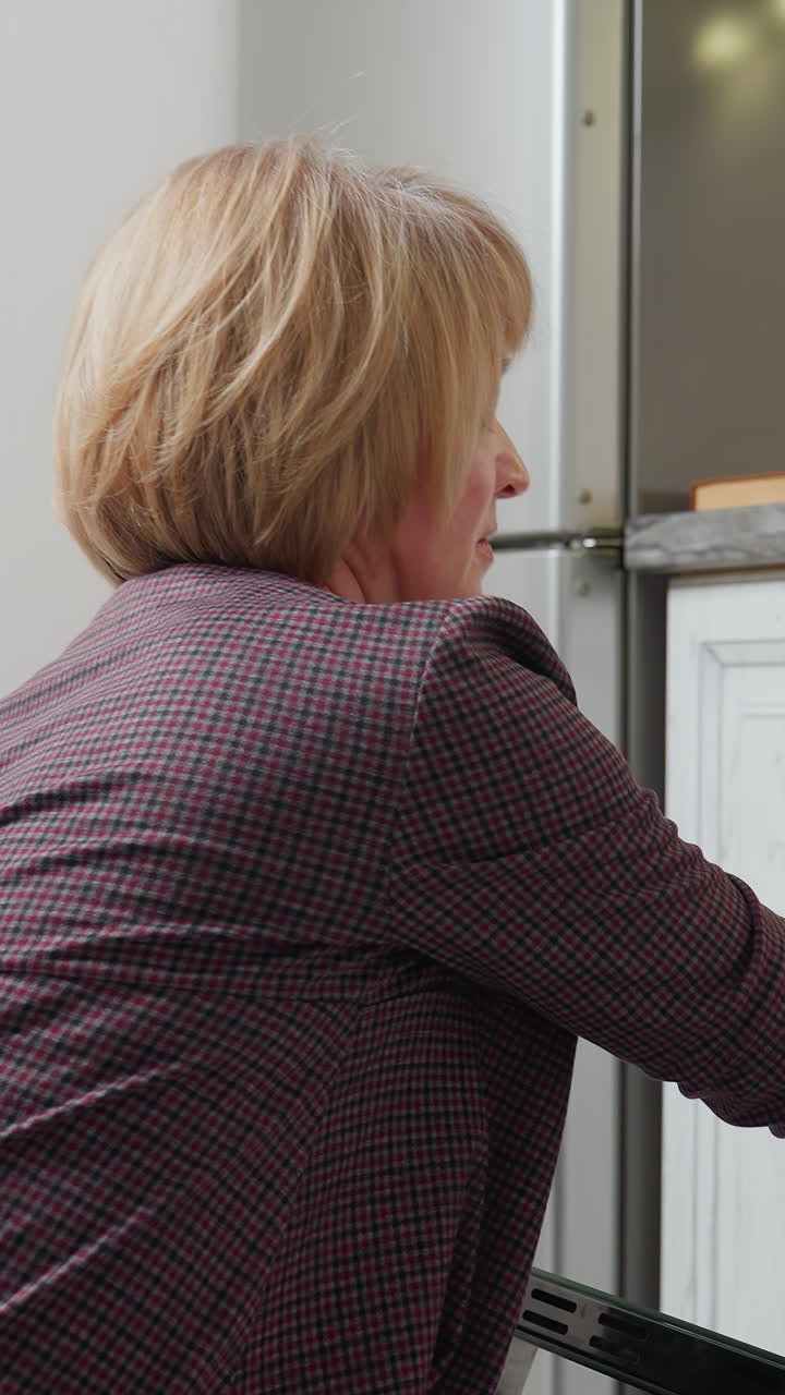 중년 여성은 오븐 문을 열고, 트레이에 쿠키를 넣고, 냉장고에 서서 현대 부에서 빛을 반사하고, 쾌적하고 잘 조명된 부 환경에서 은 상품을 준비합니다.