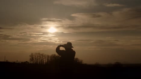silhouette of adult in hat stands to take picture of golden sunset, wide