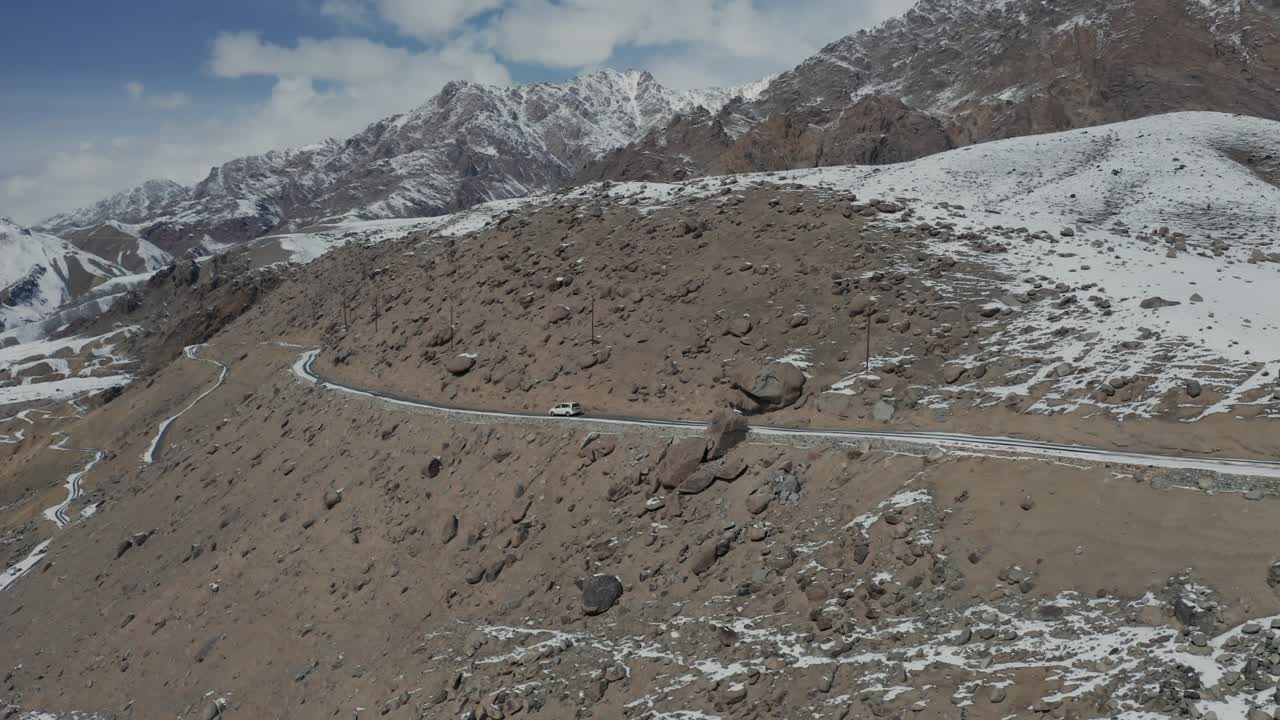 Vehicle traveling on curved highway through high-altitude terrain in Ladakh with snow patches and brown hillsides. Descending drone tracking shot follows the road through the Himalayan landscape