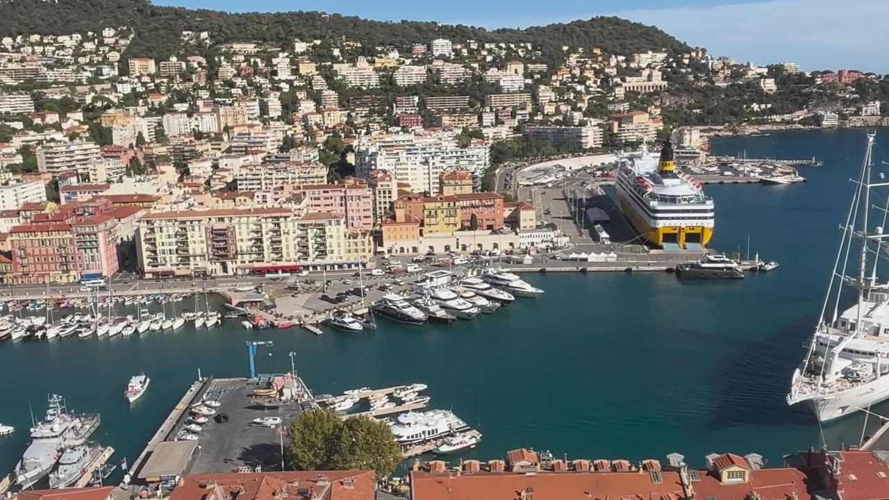 Vista elevata sul Porto di Nizza con yacht di lusso, una nave passeggeri ormeggiata, edifici colorati sul lungomare e case in collina sulla Costa Azzurra
