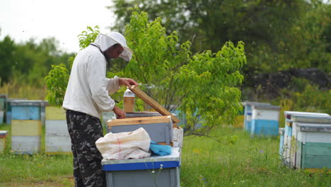 Beekeeper works at apiary. Man in protective clothes checks beehives and honeycombs. Apiculture concept. Video from side.