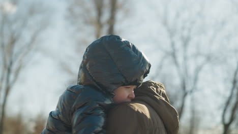 a back view of a father carrying his child while walking, the father is dressed in a brown jacket and beanie, while the child is snug in a black puffy jacket, with blurred the trees and sky