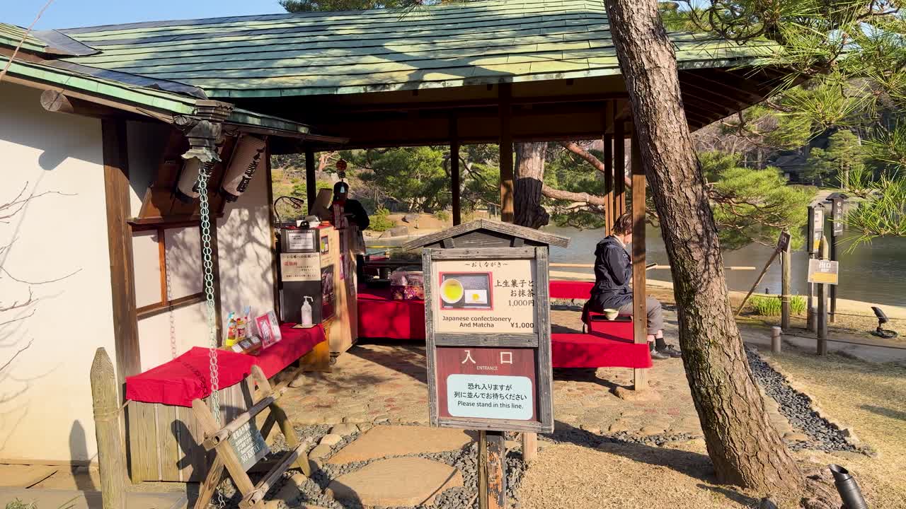 Statische Weitwinkelaufnahme eines hölzernen Teehauses mit roten Bänken in einem sonnigen japanischen Garten.