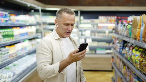 A-middle-aged-man-walks-through-a-grocery-store-talking-on-a-video-link.-Headphones-in-the-ears.-Shopping-trip.-Modern