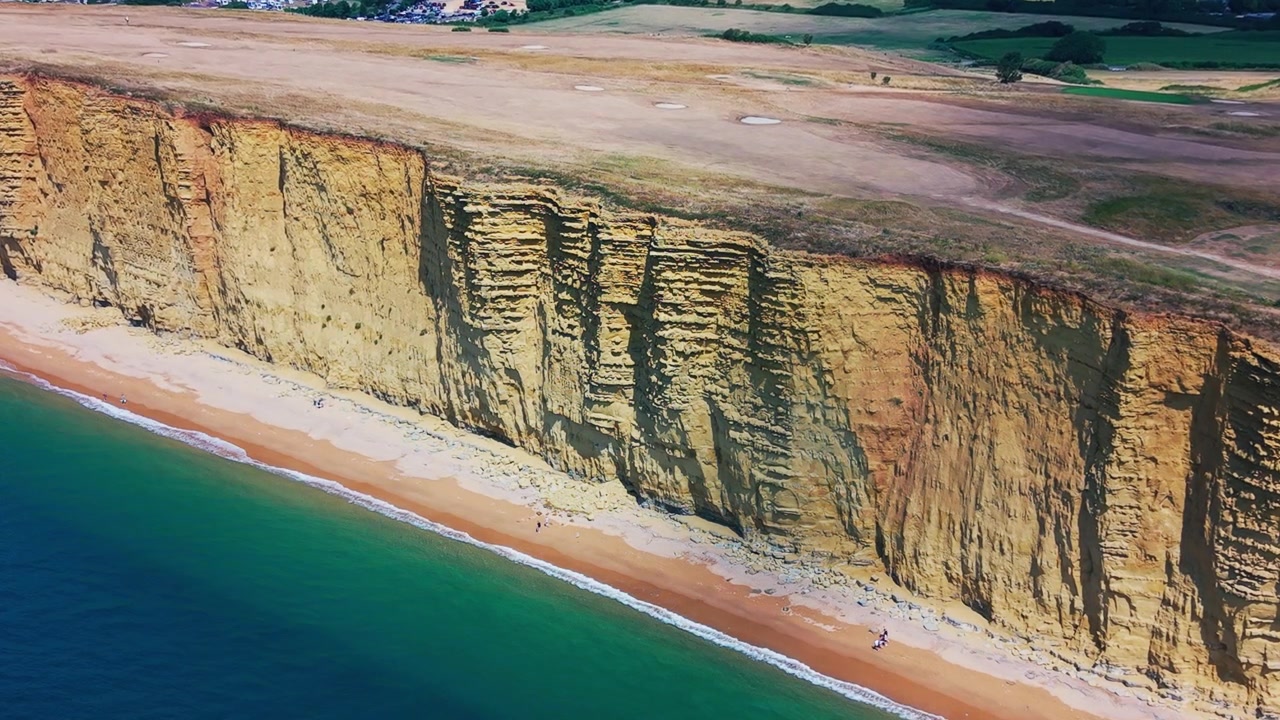 Rekaman Drone Udara Detail Bergerak di Sepanjang Tebing Berlapis Menjulang Tinggi di West Bay, Dorset, Inggris. Pemandangan Ini Menampilkan Formasi Geologi yang Terekspos, Pantai Sempit, dan Laut Pirus.