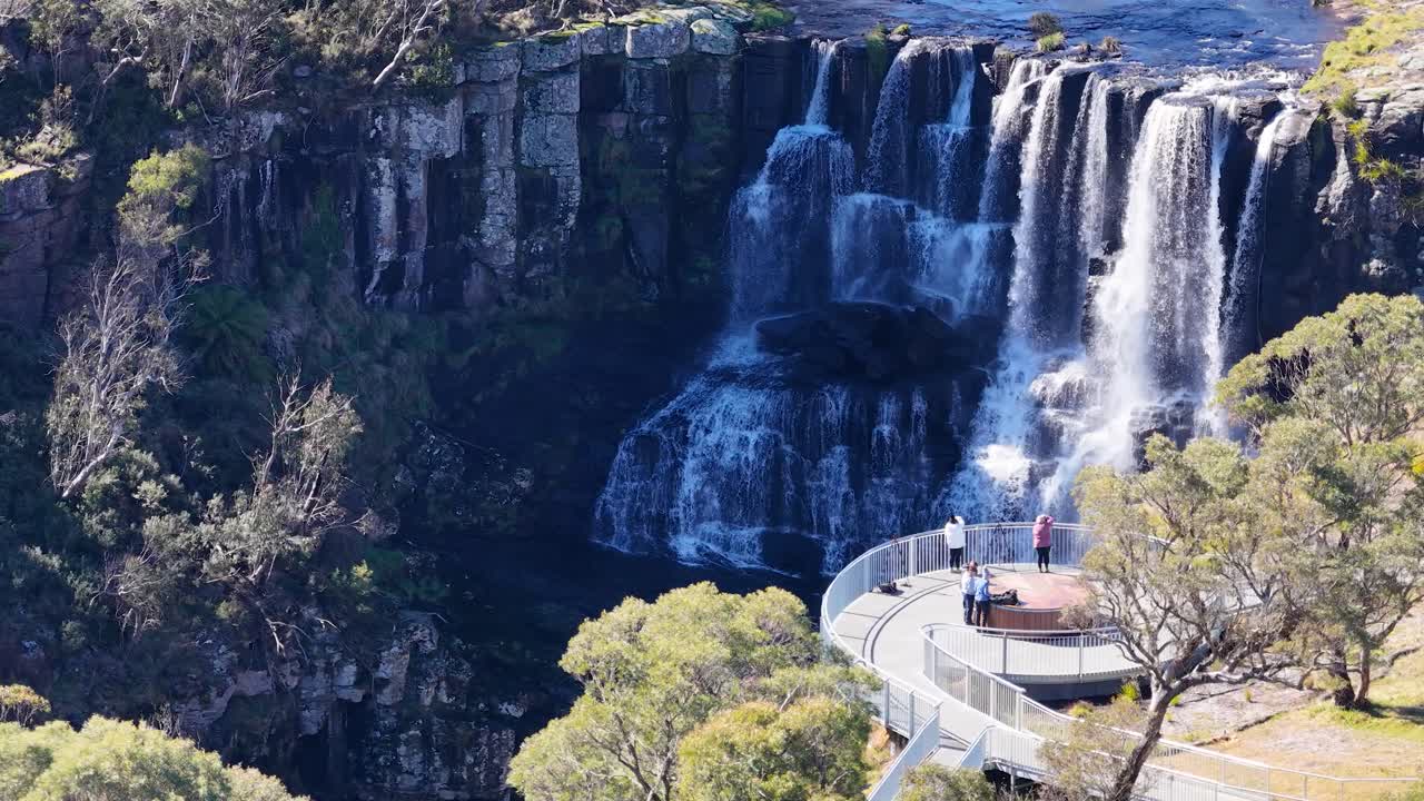 Tourists stand on a curved clifftop viewing platform overlooking a multi-tiered waterfall, surrounded by lush trees, captured in bright daylight with steady aerial camera movement