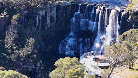 tourists stand on a curved clifftop viewing platform overlooking a multi-tiered waterfall, surrounded by lush trees, captured in bright daylight with steady aerial camera movement