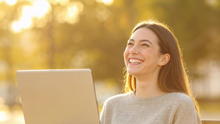 Donna sorridente che indossa un maglione grigio chiaro, ride leggendo un messaggio divertente su un laptop argentato al parco