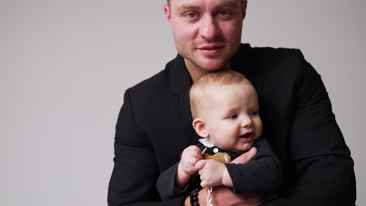 Caucasian male wearing black jacket embracing a cute little baby. Loving father kisses his beloved baby on the head. White backdrop.