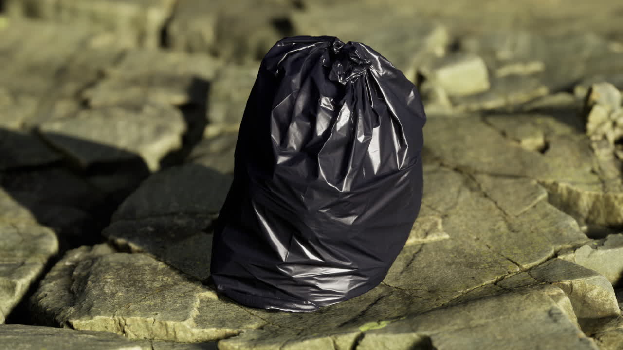 Black trash bag rests on rocky terrain near a water source during daylight