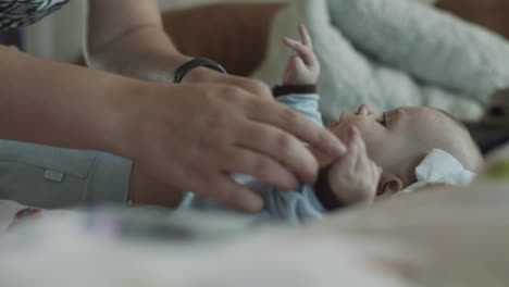 Young-mother-finishing-dressing-up-her-baby-boy-infant,-who-lies-on-the-coach-with-colorful-bedsheet