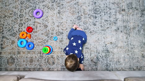 tiny kid sitting on the floor puts the phone under sofa. toy pyramid on the floor near the baby. view from top.