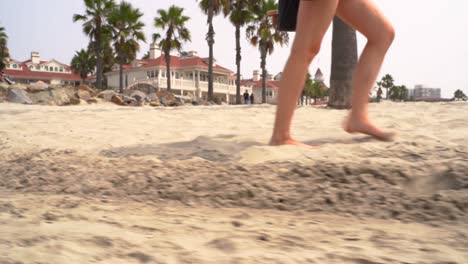 Low-shot-of-female-legs-walking-down-to-the-beach-on-golden-sand-with-palm-trees-and-resort-in-the-background