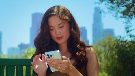 portrait of happy lady using phone in park. asian woman read message on gadget.